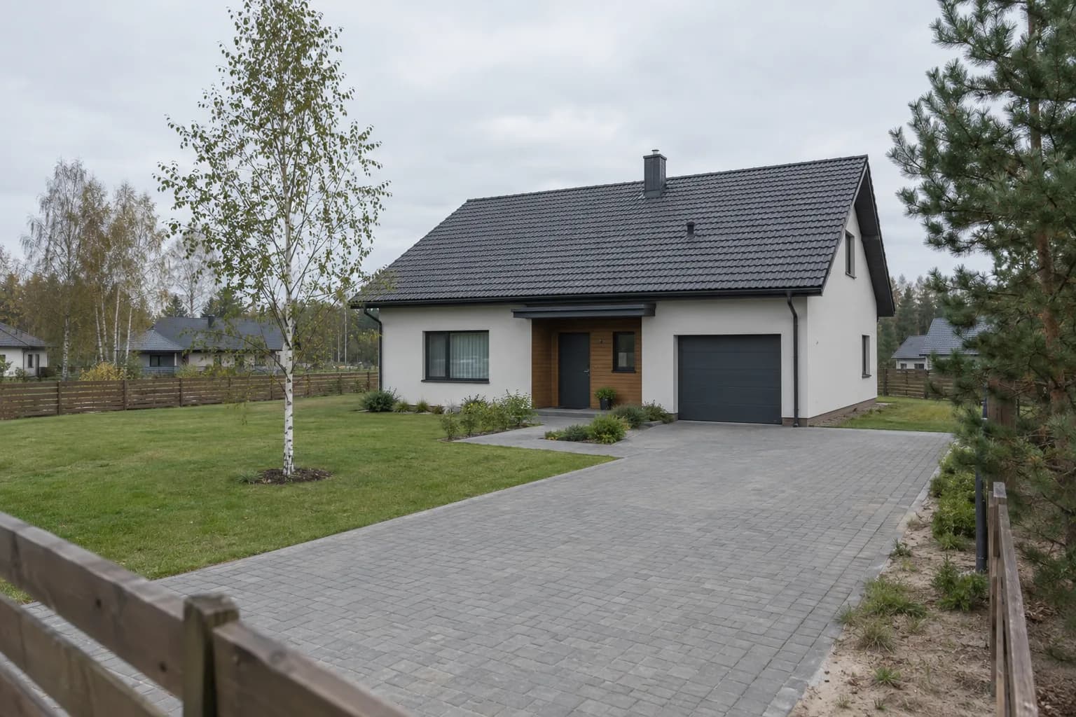 Namas su betoninių trinkelių įvažiavimu ir veja, šalia auga beržas ir pušis
